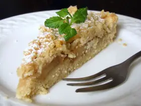 Pyszna tarta z jabłkami i kruszonką, oprószona cukrem pudrem, z listkiem mięty. Obok widelczyk.