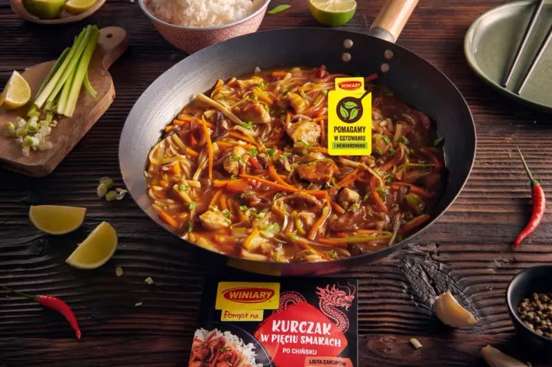 Kurczak w pięciu smakach po chińsku w woku, z warzywami i ryżem w tle. Obok limonki, chili i czosnek.